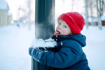 Child in winter jacket and red hat licking a snow covered metal pole outdoors, cold playful moment, gloves holding pole, snowy background, exploration and childhood curiosity and smile