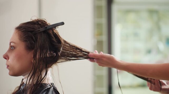 side profile wet hair being combed through, white woman client listens during stylist consultation while long damp strands are sectioned and detangled with clip and gentle comb, careful handling