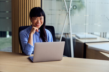 Fototapeta premium Asian Woman at Modern Office Desk With Laptop Smiling at Camera in Workspace