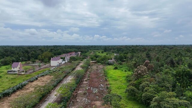 The aerial view shows a row of new houses bordering a large, undeveloped green space in a tropical region. 