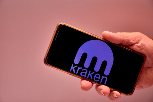Smartphone with screen on showing Kraken logo, held by a human hand in a digital trading scene.