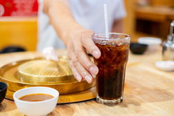 Close-up of a hand reaching for a cold iced drink glass on a wooden table, next to brass Mookata or Asian BBQ grill plate. Dinner, dining, and refreshing beverage concept at a restaurant with friends