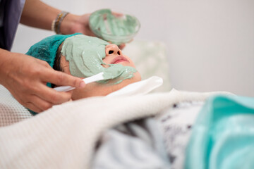 Asian woman receiving professional facial treatment at beauty spa salon. Beautician applying green natural clay mask on face with spatula. Wellness, skincare and rejuvenation concept