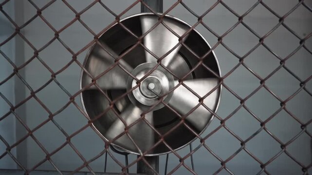 Spinning metal fan behind rusty wire fence creates industrial ventilation and cooling, hinting at confinement and machinery's relentless work