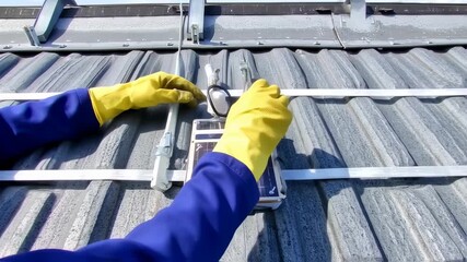 A worker in a blue outfit with yellow gloves installing a solar panel on a corrugated metal roof - Powered by Adobe