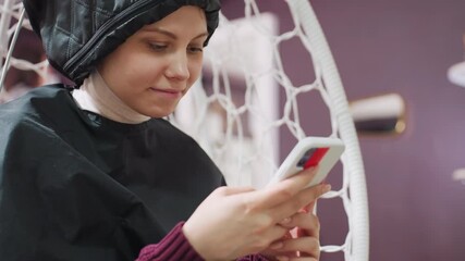 caucasian woman in hood dryer booking appointment on phone, focused expression, salon shelves in background, hanging chair comfort, scheduling via mobile app, waiting during treatment, tidy modern