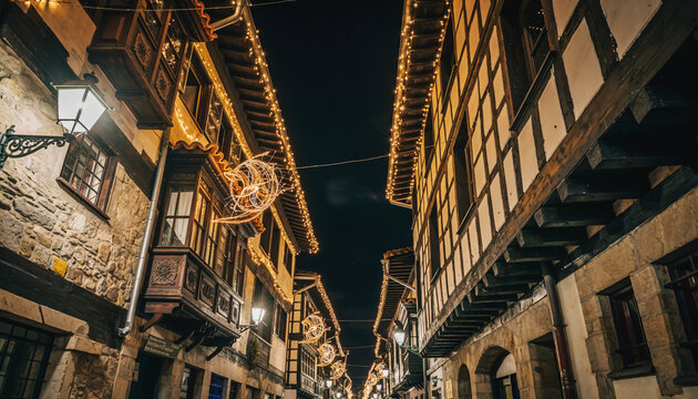 calle estrecha de un casco antiguo, iluminada con luces navide&ntilde;as c&aacute;lidas que bordean los tejados y balcones de edificios de piedra y madera, creando un ambiente festivo y acogedor durante la noche.