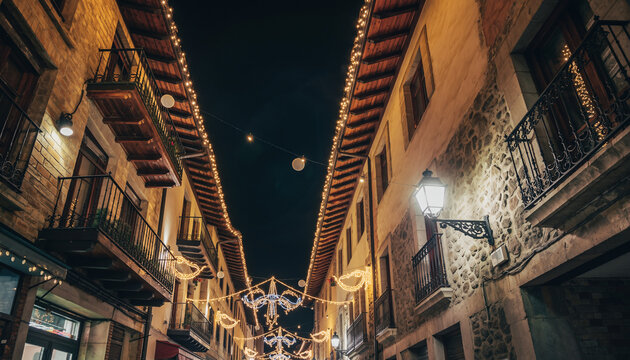 calle estrecha de un casco antiguo, iluminada con luces navide&ntilde;as c&aacute;lidas que bordean los tejados y balcones de edificios de piedra y madera, creando un ambiente festivo y acogedor durante la noche.