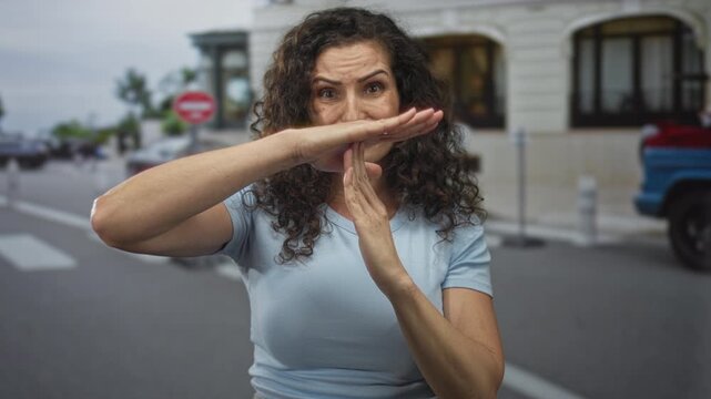 Curly haired hispanic woman wearing light blue shirt extends hands in timeout gesture on busy city street; pause warning.