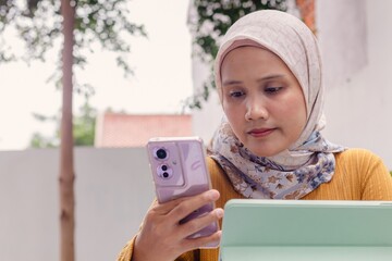 A young Asian Muslim woman working remotely using a tablet and smartphone at an outdoors coffee shop