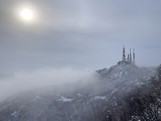 Fototapeta premium Snowy Mountain Peak in Winter Fog with Bare Trees and Communication Towers 
