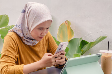 A young Asian Muslim woman replying to messages on her smartphone at a coffee shop. Outdoors.
