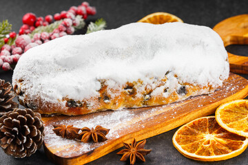 Classic Christmas Stollen covered with powdered sugar icing on wooden board with dried oranges, pine branches and cones on black textured surface. Traditional holiday food, sweet fruit loaf