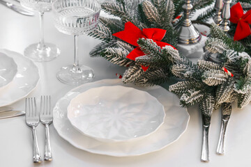 Bright Christmas table setting with white plates, crystal glasses, silver cutlery, and frosted pine branches decorated with red bows, creating an aesthetic, elegant winter holidays atmosphere.