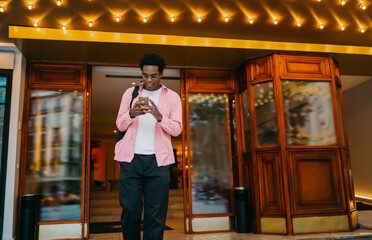 Smiling Black man reading phone while exiting hotel, dressed casually, reflecting relaxed mobile routine in dynamic city setting