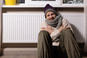 freezing man with hat and scarf in front of radiator in cold room at home. heating problems