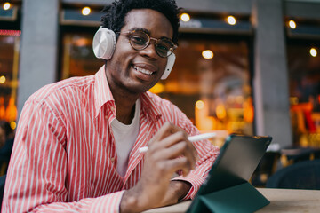 Obraz premium Young African man smiling with stylus in hand, using tablet outdoors with headphones, reflecting positivity, creativity and modern freelance energy in tech workspace.