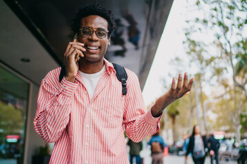 Cheerful African freelancer in striped shirt talking on phone with expressive gesture, standing on urban street, showing energy, connection and remote work flexibility.