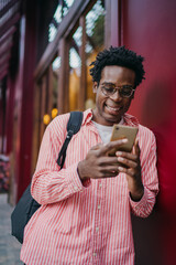 African man with glasses smiling at phone, standing against red wall, highlighting digital fluency, freelance autonomy and mobile-first tech engagement in city setting.