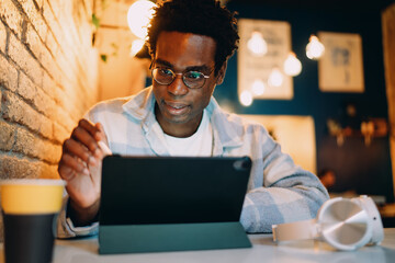 Smiling young man engaged with digital tablet using stylus in a warmly lit café. Scene reflects creativity, modern digital tools, remote work flexibility, and casual productivity mindset.