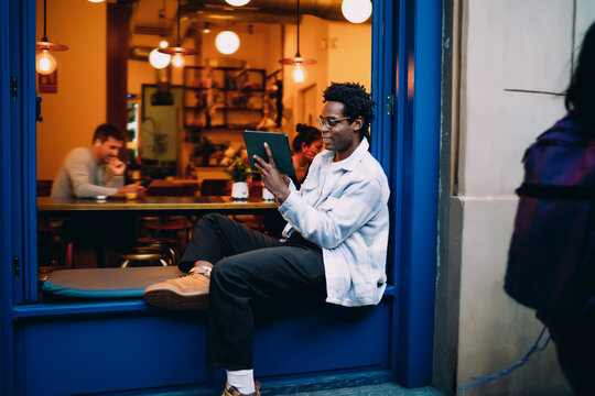 Full-body shot of man using tablet while seated in window ledge, blending physical rest and digital activity in a creative urban lifestyle expression.
