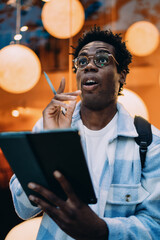 Young man looks upward in realization while holding a tablet and stylus. Tech interaction captured in a spontaneous moment of insight, embodying mobile thinking and digital expression. © BullRun