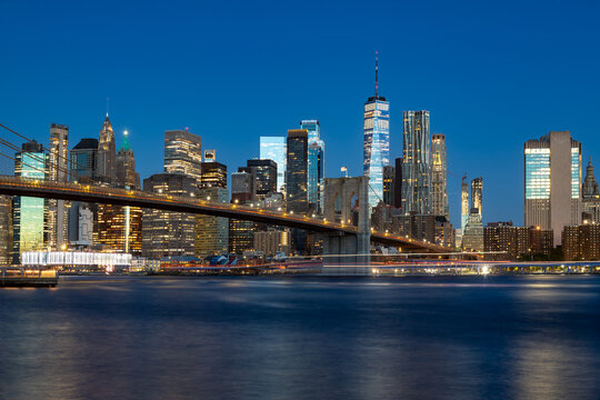 A panoramic view of the New York City waterfront at dawn, featuring the brilliantly lit Downtown skyline and the historic Brooklyn Bridge.