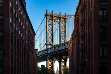 The majestic Manhattan Bridge seen from the street level in the DUMBO neighborhood of Brooklyn, framed by classic red brick buildings.