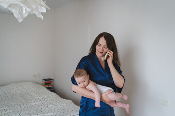 A woman in a blue robe stands in a well-lit bedroom, holding her baby in one arm while speaking on the phone. The room features a bed with white sheets and a light-colored decor.
