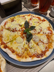 A sliced thin-crust pizza topped with melted mozzarella cheese, tomato sauce, and fresh basil is served on a round blue ceramic plate. The Italian dish sits on a light wooden table surface in a restau