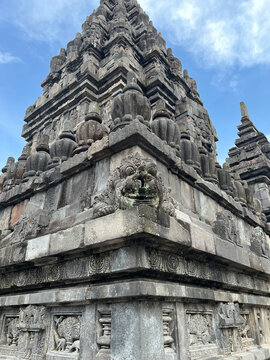 A low-angle view captures the grey volcanic stone structure of an ancient Javanese Hindu temple. A protruding Kala face sculpture dominates the corner, with tiered decorative ratna motifs above.
