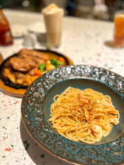 A blue, textured ceramic bowl holds spaghetti with prawns, olive oil, and fried shallots in the foreground. A sizzling steak platter and a milkshake are visible in the shallow, blurred background.