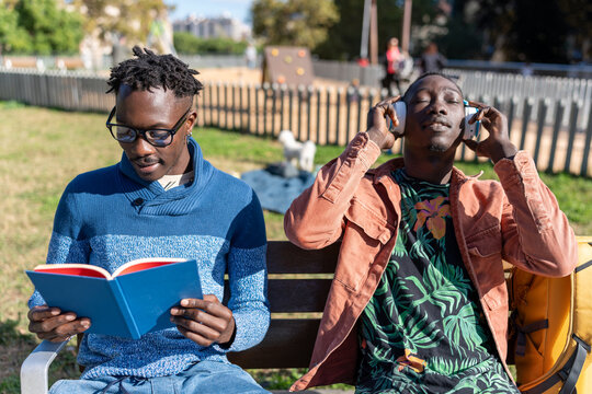 Friends relaxing outdoors, one reading a book, other listening to music