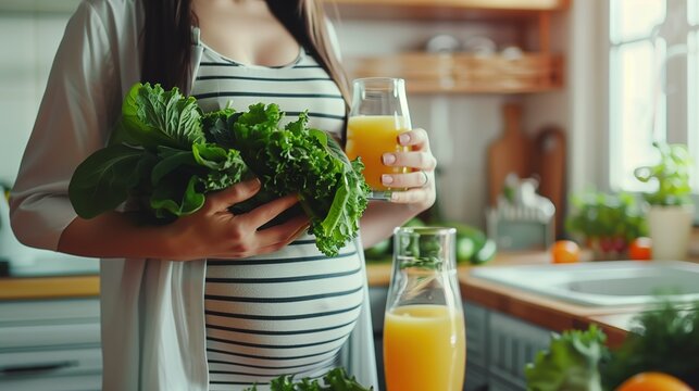 National Folic Acid Awareness Week: Pregnant woman holding fresh green leafy vegetables and orange juice in modern bright kitchen