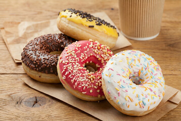 Four donuts with yellow, chocolate, pink, and white toppings resting on brown paper beside a coffee cup on a wooden table