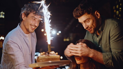 Friends bringing cake sparkler candle to happy woman in restaurant closeup.
