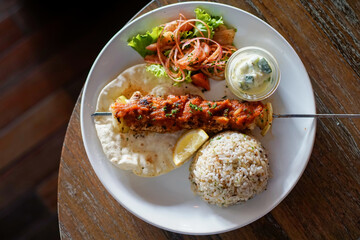Overhead shot of grilled chicken kebab with rice, flatbread, fresh salad, lemon, and yogurt sauce...
