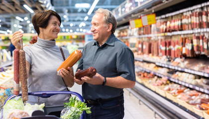 Mature couple diligently choosing tasty sausage in supermarket