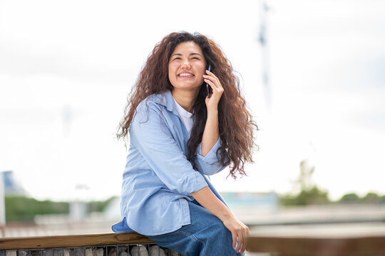 Smiling Asian woman sitting outdoors on a sunny day and talking on her smartphone