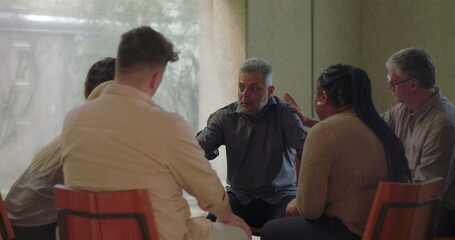 Man receives emotional support from group therapy members, seated in circle as participants place...