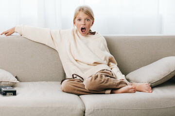 A young girl sits on a light sofa wearing a cozy sweater, her wide eyes and open mouth capturing a...
