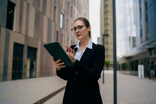 Businesswoman checks tablet while standing outdoors with city architecture around her, conveying digital access, mobile executive workflow and tech‑first business mindset. - Powered by Adobe
