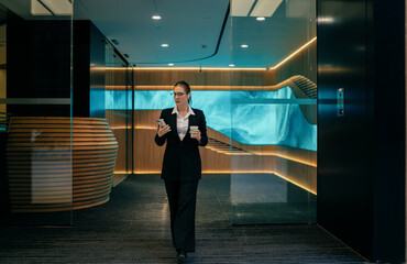 Caucasian businesswoman in suit walking in modern office lobby, checking smartphone and holding coffee, multitasking during busy workday with focus and digital efficiency