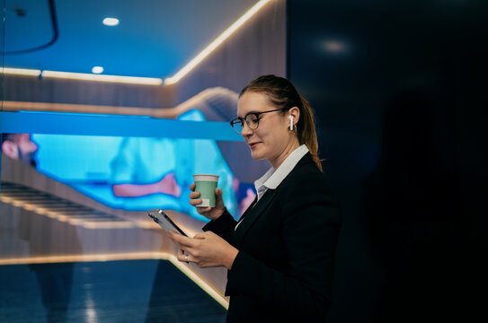 Caucasian woman walking in tech-inspired office, looking at smartphone while holding coffee, multitasking in sleek business environment, focused on digital work and communication - Powered by Adobe