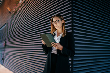Focused businesswoman standing in front of striped wall, immersed in reading tablet screen, reflecting digital immersion and solo work ethic