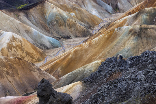 View of vibrant, undulating hills painted in earth tones and cool blues, dissected by a winding stream, contrasted by dark volcanic rock, Landmannalaugar, Rangarbing ytra, Iceland.