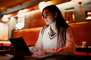 Woman in glasses working on a tablet in ambient lighting, showcasing technology in contemporary life, highlighting connection between focus, digital access, and creative tasks.