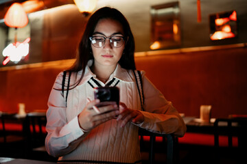 Front view of young woman in glasses using a smartphone with stylus, focused on screen under ambient lighting, illustrating tech fluency, digital identity, and mobile-first behavior.