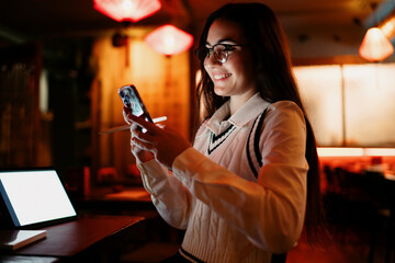 Smiling woman holding a smartphone in one hand and tablet under arm, capturing a moment of mobile communication and tech‑enabled social interaction in ambient lighting.