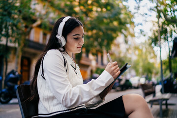 Focused young woman wearing headphones works on tablet with stylus outdoors, representing mobile productivity — blending portable technology with creative concentration.